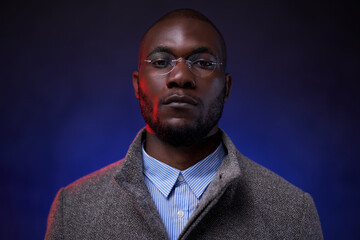 Stylish African American man in gray jacket and glasses on dark background with blue light. Studio shot