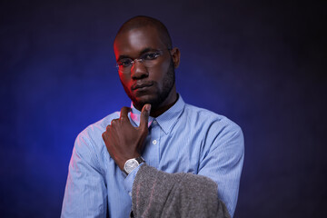 African American businessman in blue shirt and glasses, looks pensive and looking on camera. Studio portrait on a dark background with blue light