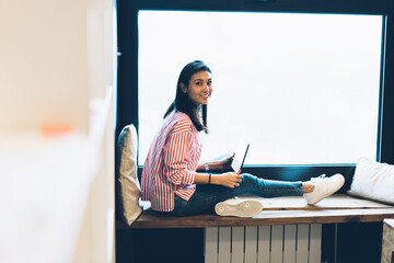 Full length portrait of gorgeous cheerful brunette student waiting for useful application installation on modern netbook during sitting against window with advertising area for promotional content