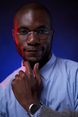 African American student in blue shirt and glasses, holds hand on chin and smiling. Studio portrait on a dark background with blue light