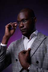 Portrait of a African American businessman in glasses and stylish checkered jacket on a dark background illuminated by purple light. Vertical photo