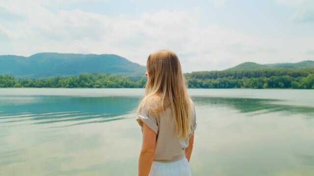 Portrait of young beautiful woman in casual clothes, authentic and natural beauty look at amazing nature landscape of mountains and lake, turn around and look into camera. Wind blows hair, deep eyes