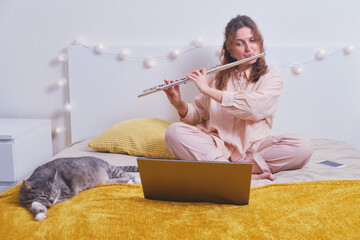 Young woman plays the flute on the bed with a cat, online music lessons. Girl with a musical...