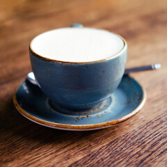 Cup of coffee with milk foam, saucer and spoon on a wooden table of a summer cafe, close up