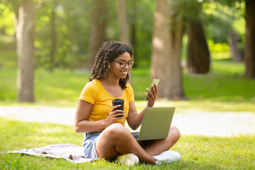 Cool black girl with coffee, smartphone and laptop computer studying online at summer park
