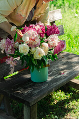 Woman arranging bouquet of peony flowers in milk can on wooden garden bench