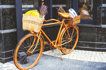 Fototapeta na wymiar Orange retro bike with knitted straw basket with cheese head and bouquet of dry lavender inside attached to handlebars. Concept of natural eco materials, shopping, plastic free, zero wast.