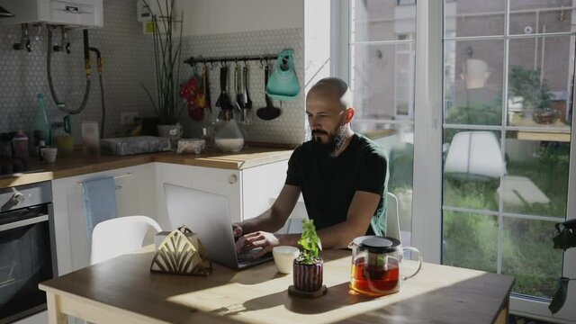 Freelancer programmer working from home on kitchen and using laptop. Bearded man working with a computer, prints text on keyboard. Self entrepreneur sitting and working at his modern apartment.