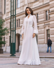 Elegant fashionable stylish female model in long white dress walking at city street. Outdoor full...