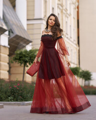 Elegant fashionable female model in red long evening dress with open shoulders and sleeves standing and posing at city street. Beautiful caucaisan woman with wavy hair and makeup outdoors portrait