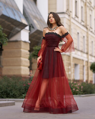 Elegant fashionable female model in red long evening dress with open shoulders and sleeves standing and posing at city street. Beautiful caucaisan woman with wavy hair and makeup outdoors portrait