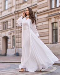 Fototapeta premium Elegant fashionable stylish female model in long white dress walking at city street. Outdoor full length portrait. Caucasian lady with wavuy hair and makeup