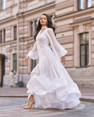 Elegant fashionable stylish female model in long white dress walking at city street. Outdoor full length portrait. Caucasian lady with wavuy hair and makeup