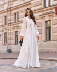 Elegant fashionable stylish female model in long white dress walking at city street. Outdoor full length portrait. Caucasian lady with wavuy hair and makeup