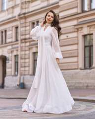 Obraz premium Elegant fashionable stylish female model in long white dress walking at city street. Outdoor full length portrait. Caucasian lady with wavuy hair and makeup