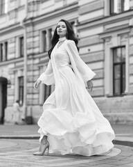 Elegant fashionable stylish female model in long white dress walking at city street. Outdoor full length portrait. Caucasian lady with wavuy hair and makeup