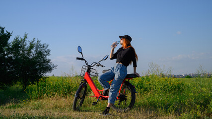 Fototapeta na wymiar Caucasian girl stand near red electro bicycle at background of blue sky drinking water from bottle, resting, enjoy after bike ride at sunny summer day. fitness, sport, people healthy lifestyle concept