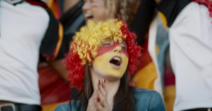 Young woman with german flag color wig and face paint clapping and chanting with group of fans for their national team. German football team supporters cheering during a match at stadium.
