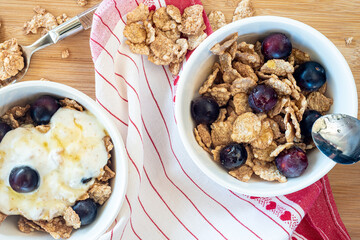 Bowl for breakfast with cereals, oatmeal, berries, honey and yogurt, on a wooden cutting board with a tea towel and teaspoons.