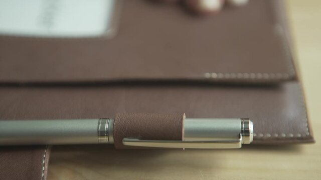 Close up shot of man hand putting silver ball pen into pen holder on light brown leather note pad bullet journal planner