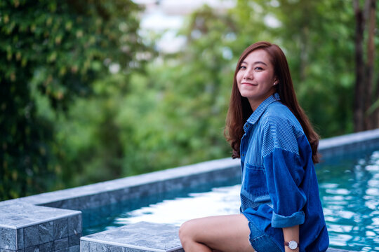 Portrait image of a beautiful young asian woman enjoyed sitting by swimming pool with green nature background