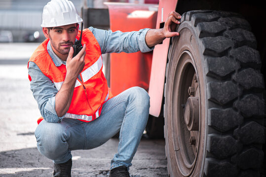 Engineer or mechanic man checking and maintenance forklift wheel tyre at container box cargo and using walkies talkie call with his supporter to confirm inspect before use.