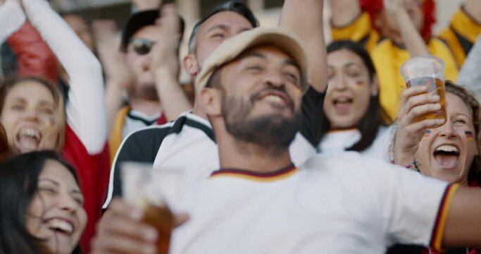 Crowd of spectators cheering at sports event, man holding a glass of beer. Germany football team supporters actively jumping and chanting in crowd.
