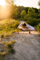 Young woman with stretching body practicing yoga and performing Eight-Angle Pose outside in park evening on background of sunlight. Female doing advanced yoga exercises outdoors at green grass.