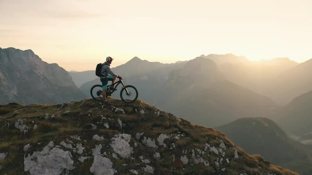 Aerial - Flyover shot of a mountain biker standing on top of the mountain at sunset. 4K