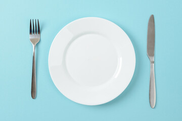 Empty white dinner plate, table knife and fork on a blue background. Set table. Copy space on dish for food design.