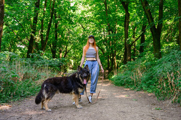 caucasian girl with long red hair is walking with her big dog, summer sunny day, green forest, woman is wearing a blue jeans, white top and black baseball cap. pet, domestic animal and people concept