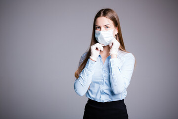 Girl with long hair and gloves straightens a medical mask on her face and looks at the camera on a gray background