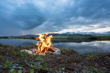 Bonfire next to lake at evening during blue hour with mountain scenery view.
