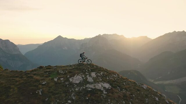 Aerial - Flying backwards over mountain biker resting on his bike on top of the mountain peak at sunset. 4K