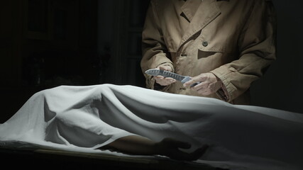 Maniac killer in a cloak stands over a victim covered with a white cloth with a knife in a dark room