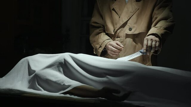 A maniac killer in a coat holding a knife stands near a man on a table covered with a white cloth in a dark room, the criminal is going to cut the victim