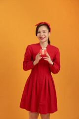 Portrait of a happy pretty asian woman holding present box while standing and looking at camera isolated over orange background