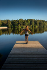 Fototapeta na wymiar Young woman with outstretched arms standing relaxing or stretching on wooden pontoon staring at lake. evening light Back view .