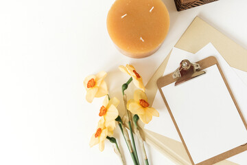 Blank paper sheet clipboard tablet pad with mock up copy space. Beautiful home office table desk workspace with fresh narcissus flowers, wooden casket, candle, albums. Flat lay, top view art concept