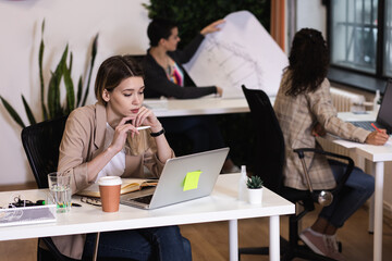 Portrait of a young female freelancer working in an office while sitting at a table with colleagues in the background.