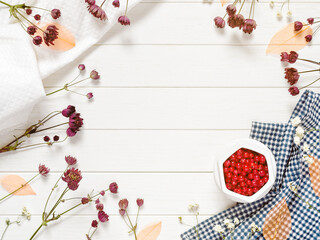Layout with red currant berries in a white bowl. The mockup is decorated with a towel and dried flowers. White wooden background. Flat lay. Top view. Copy space.