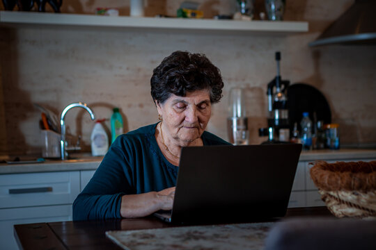 An Older Senior Woman With Black Short Hair Is Sitting In The Dining Room And Using A Laptop Computer. She Is Looking For Some New Recipes For Cooking Online.