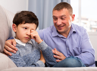 Young father consoling preteen son sitting at sofa after quarrel at home