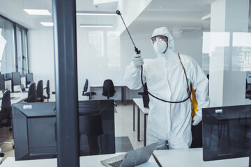 Office disinfection during COVID-19 pandemic. Man in protective suit and face mask spraying for...