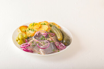 Fototapeta na wymiar Slices of pickled herring with red onion, fermented cucumber, fried potatoes, olives and herbs on a light wooden table. Copy space
