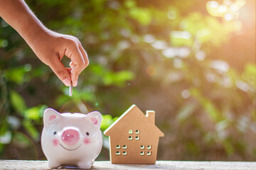 Hand holding and putting coin into piggy bank, Toy wooden house on table with natural blurred...
