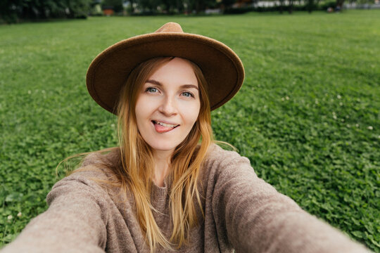Beautiful young hipster woman in hat taking selfie on green grass at the park. Looking at camera and show tongue. Travel and active life concept. Outdoors