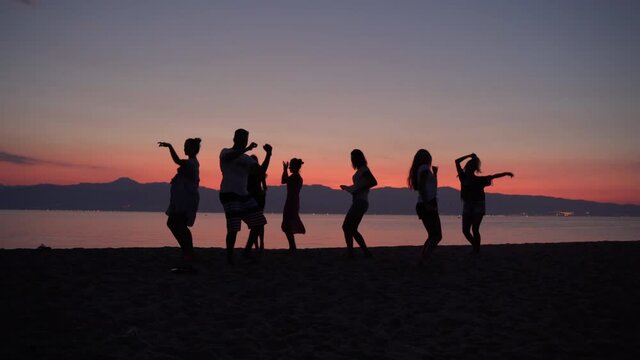 Silhouettes of cheerful dancing people on beautiful sea sunset background. Friends dancing and jumping, beach party and disco music. Fresh summer beverages for relax and holidays enjoyment