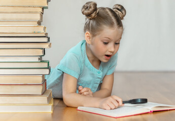 A little blonde schoolgirl looks at book through a magnifying glass in surprise, tearing her eyes off. A pile of books on blurred light background. Back to school. Education. Free time for the child.