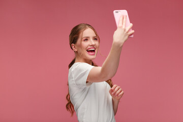 Funny young redhead woman making selfie. Smiling girl wearing white t-shirt holding pink smartphone, making faces on camera, posing for selfie isolated on pink background.